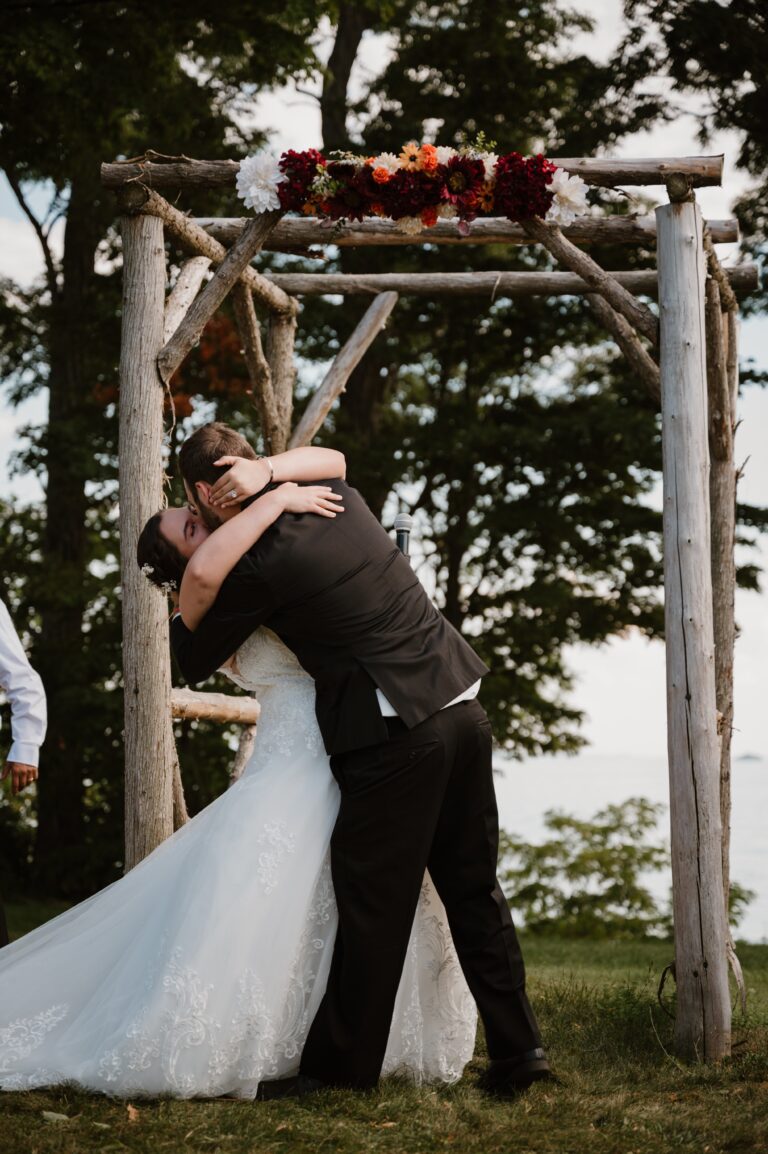 couple kissing at a vineyard wedding below a wooden arch with lake champlain in background